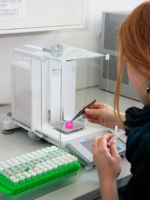 The photo shows a laboratory at the Institute of Geography. A woman with long red hair is only half visible on the right-hand side of the picture. She is sitting at a piece of laboratory equipment and is just putting a sample into the measuring device. In front of her at the table is a container holding various samples.