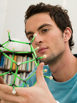 The photo shows a close-up of a young man with short black hair and a dark shadow of a beard. He is wearing a turquoise T-shirt and is looking intently at his hand, in which he is holding a model of a molecular structure. A shelf with books can be seen in the background.